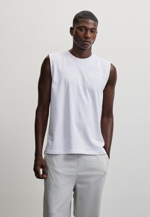 Young man with dark skin wearing a white sleeveless shirt and light gray pants standing against a plain light background.