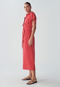 Koraloowa sukienka koszulowa z krótkimi rękawami, dwoma przednimi kieszeniami, paskiem w talii i prostym dołem. Noszona z czarnymi płaskimi sandałami.