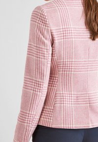 Veste à carreaux rose avec un design ajusté, un col à revers et un tissu texturé, mettant en avant des carreaux contrastants de différentes tailles.