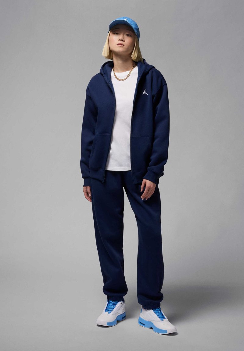 Joven con chándal azul marino, camiseta blanca, gorra azul, cadena de oro y zapatillas blanco-azul, de pie contra un fondo gris liso.
