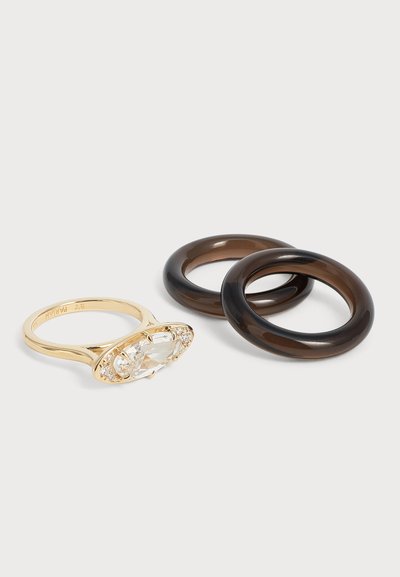 Bague en or avec une grande pierre ovale claire et deux anneaux ronds translucides foncés sur un fond clair.