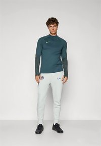 Long-sleeve teal athletic top with black textured sleeves, paired with light gray pants featuring blue Nike logo, worn with black shoes.