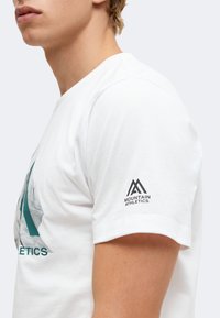 Πλάγια όψη ενός ατόμου που φοράει ένα λευκό T-shirt με λογότυπο "Mountain Athletics" στο μανίκι και μερικό γραφικό στο στήθος.