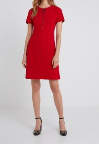 Robe rouge ajustée à manches courtes, encolure ronde, détail de fermeture éclair à l'avant, et légère évasée à l'ourlet. Portée avec des chaussures à talons hauts noires cloutées.