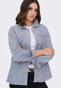 Noor naine, kellel on õlavarrest lõikuvad pruunid juuksed, kandes helesinist sametist jopet valge toppi ja mustade pükste kohal, käed ristatud.