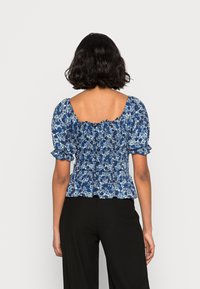 Blå blommig blus med puffärmar och smockad midja samt fyrkantig halsringning, med ett strukturerat mönster på en mörk bakgrund. Svarta vida byxor.