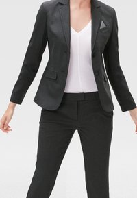 Blazer su misura grigio scuro con due bottoni, rever a scollo e taschino sul petto. Abbinato a pantaloni su misura dello stesso tessuto e a un top bianco con scollo a V.