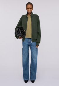 Tmavě zelená bombera s vysokým límcem, olivově zelený top, široké modré džíny a černá skládací kabelka, nošeno s černými špičatými botami.