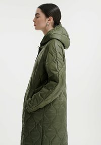 Cappotto lungo imbottito in verde oliva, con cappuccio, texture trapuntata, tasche laterali e silhouette slanciata.