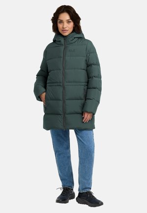Donna che indossa un cappotto imbottito scuro con cappuccio, jeans blu e sneaker nere in piedi davanti a uno sfondo chiaro e uniforme.