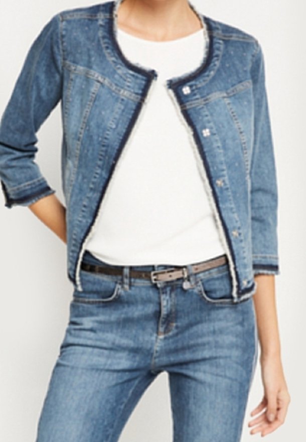 Femme portant une veste en jean bleue aux bords effilochés, une chemise blanche, un jean bleu et une ceinture marron, debout avec une main derrière le dos.