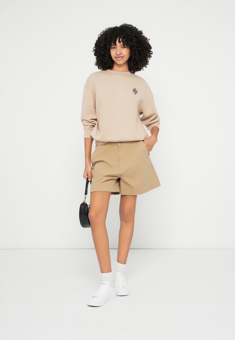 Beige gebreide trui met een klein zwart geometrisch logo, gecombineerd met khaki shorts. Witte sneakers en een zwarte handtas maken de look compleet.