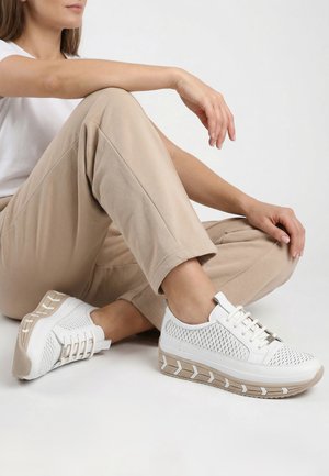 Woman seated with legs crossed wearing beige pants and white sneakers with woven texture and light brown patterned soles.