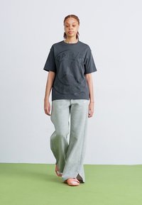 Zwart oversized T-shirt met een rond logo en tekst, gecombineerd met lichtgrijze wijde sweatpants en oranje slippers op een groene vloer.