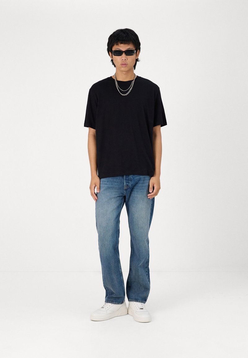 Camiseta negra de manga corta, jeans de denim azul, zapatillas blancas, cadenas de plata en capas y gafas de sol oscuras. Estilo sencillo y casual.