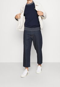Maglione a girocollo blu navy con accenti a righe bianche, giacca in denim color crema, pantaloni larghi scuri e sneakers bianche con accenti beige e verdi.