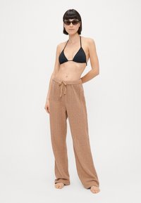 Μαύρο μπικίνι τοπ και ανοιχτό καφέ, υφασμένα παντελόνια σε φαρδιά γραμμή με πλέξη και μέση που ρυθμίζεται με κορδόνι. Μοντέλο που φοράει γυαλιά ηλίου.