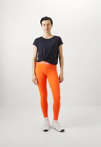 Černý krátký top s uzlem v pase, doplněný jasně oranžovými legínami s bílým páskem s logem a bílými teniskami.