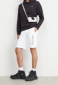 Zwarte hoodie met logo, witte shorts met frontprint en zwarte chunky sneakers met witte accenten. Schoudertas in wit en zwart.