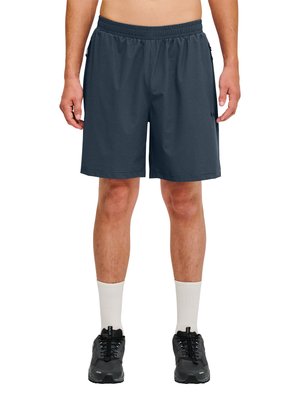 Mand iført mørkegrå atletikshorts, hvide crew-sokker og sorte løbesko, stående med armene afslappet ved siderne.