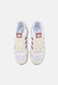 White and cream sneakers with mesh and suede textures, featuring three brown stripes and a branded tongue logo. Flat laces.
