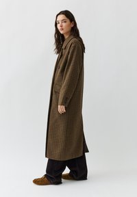 Langer, brauner karierter Mantel mit notch-Kragen, zwei vorderen Taschen und einem einzigen Knopfverschluss. Getragen über dunklen Hosen und braunen Mokassins.