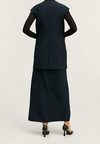 Chaleco largo sin mangas azul marino sobre una falda a juego hasta el suelo, con líneas limpias y un ajuste entallado, combinado con zapatos de tacón negro.
