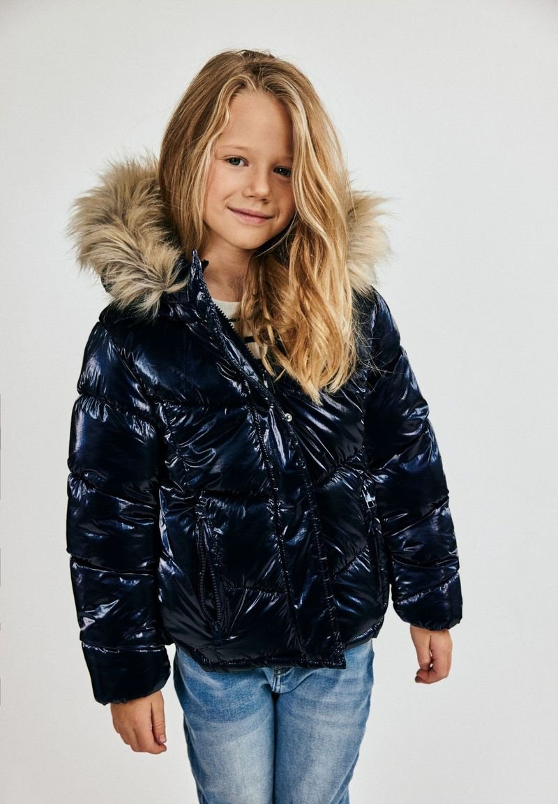 Mornarska pufasta jakna sjajnog izgleda, s kapuljačom obloženom krznom, prednjim zatvaračem i elastičnim manžetama, u kombinaciji s svijetloplavim trapericama.
