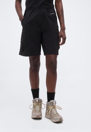 Osoba nosiaca čierne cargo šortky s elastickým pásom a béžové turistické topánky s čiernymi ponožkami, stojaca pred jednoliatym pozadím.