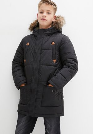 Parka noire rembourrée avec capuche en fourrure, dotée d'accents en cuir marron et de poches latérales. Texture lisse avec un design matelassé.