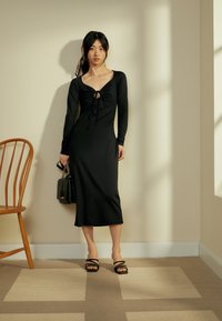 Vestido preto de manga longa com frente franzida e detalhe em fenda, corpete justo e saia fluida. Combinado com saltos strappy pretos e uma bolsa pequena.