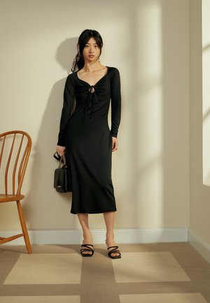 Vestido preto de manga longa com frente franzida e detalhe em fenda, corpete justo e saia fluida. Combinado com saltos strappy pretos e uma bolsa pequena.