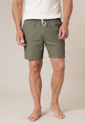 Homme portant un short texturé vert olive avec un cordon blanc et un t-shirt blanc, debout pieds nus sur un fond beige.