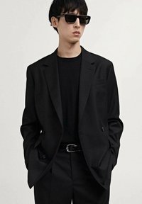 Black tailored suit with a double-breasted jacket and matching trousers, paired with a black crew-neck shirt and dark sunglasses.