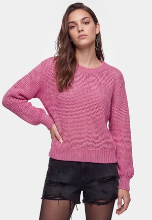Jonge vrouw met lang bruin haar, gekleed in een roze gebreide trui, zwarte gescheurde shorts, zwarte panty's en stervormige hangende oorbellen.