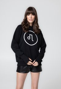 Czarna bluza z kapturem z dużym białym okrągłym logo z przodu, z prążkowanymi mankietami i dołem. Nosi się z czarnymi skórzanymi szortami.