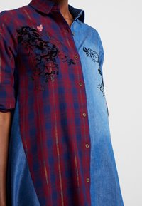 Camicia button-up con metà motivo a quadri rossi e blu e metà in denim azzurro chiaro, caratterizzata da ricami floreali neri e un piccolo dettaglio a forma di cuore.