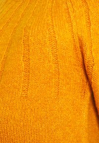 Stickad tröja i starkt gult med en texturerad yta. Har vertikala ribbade mönster och ett mjukt, mysigt utseende.