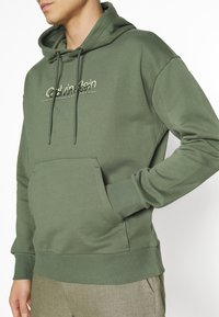 Felpa verde oliva con tasca frontale, cappuccio con cordino e polsini a coste, con un logo discreto sul petto in beige.