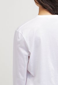Vit bomull långärmad skjorta, slät textur, avspänt snitt, rund halsringning och synliga sömdetaljer på axlarna och ryggen.