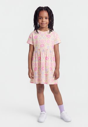 Fată tânără cu părul împletit, purtând o rochie pastel cu imprimeu floral, șosete lavandă și adidași albi, stând în picioare pe un fundal simplu.