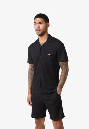 Mand med tatoveringer iført sort kortærmet skjorte med knapper og matchende shorts, stående med den ene hånd i lommen, neutral udtryk.