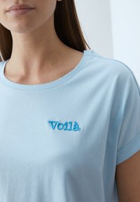 Lyseblå bomulds T-shirt med rulleærmer og en broderet "Voilà" i mørkere blå på fronten. Blød tekstur med klassisk rund hals.