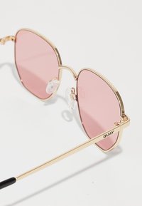 Gafas de sol color rosa con un delgado marco de metal dorado, que cuentan con almohadillas nasales transparentes y puntas de patas negras. Logo "QUAY" en el brazo.