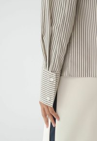 Main reposant sur le côté, portant un chemisier beige à rayures verticales noires et un poignet assorti à rayures avec deux boutons blancs.