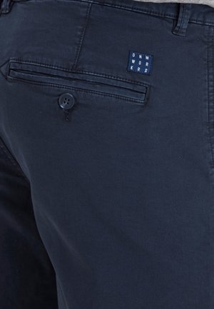 Nahaufnahme der Gesäßtasche einer marineblauen Hose mit Knopfverschluss und einem quadratischen Stofflogo mit Buchstaben in einem Raster angeordnet.