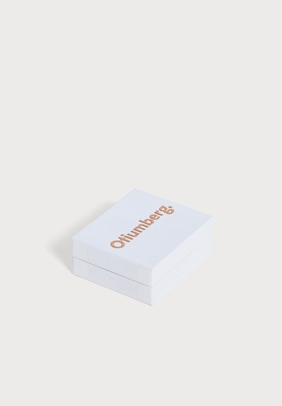Une boîte blanche à la texture marquée, portant le nom de marque « Otilimber » de couleur cuivrée, est posée sur un fond clair.