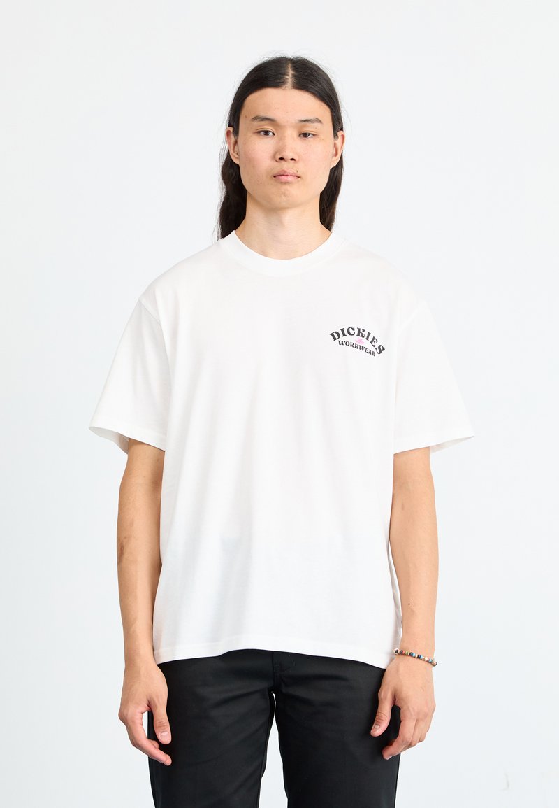 Hvit bomulls T-skjorte med korte ermer, rund halslinning, og en svart "Dickies" logo trykt på venstre bryst. Bært med svarte bukser.