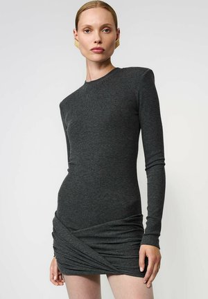 Kobieta w dopasowanej, ciemnoszarej, długosleeve mini sukience z drapowanymi detalami na spódnicy i złotymi, geometrycznymi kolczykami.