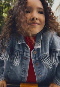 Jeansjacke mit Fransen, hellblaue Farbe, mit Knopfverschlüssen, getragen über einem roten Oberteil, ergänzt durch lockige Haare. Der Hintergrund zeigt grüne Pflanzen.